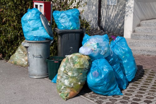 A collection of rubbish bags and waste bins situated on a paved urban driveway, with several large blue plastic bags filled with household waste placed alongside a grey metal wheelie bin, a black plastic bin, and a smaller green container. The blue bags appear tightly knotted and contain a mix of recyclable and non-recyclable waste, with some bags leaning against each other and the bins, while others rest on the ground. The background features a concrete staircase leading up to a residential building, with some greenery visible on the left side, indicating a garden or shrubbery area. The scene is well-lit with natural daylight, emphasizing the different textures such as the smooth plastic of the bags, the rough concrete of the stairs, and the metallic surfaces of the waste containers. This depiction aligns with rubbish collection and waste management services offered by Commercial Waste Basildon for local businesses or residents in the Basildon area, focusing on proper disposal and clearance of rubbish.