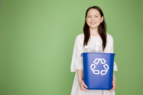 A young woman with light skin and long dark hair is standing in front of a plain green background, smiling gently while holding a blue recycling bin with a white recycling symbol on the front. She is dressed in a white, loose-fitting blouse with lace details on the sleeves. The bin is partially filled with clear plastic bottles, and she is gripping it with both hands, one on each side. The setting is simple and well-lit, emphasizing her friendly expression and the focus on the recycling bin. This scene subtly relates to waste management and rubbish reduction services available in Basildon, as represented by companies like Commercial Waste Basildon, and reflects environmentally conscious practices such as recycling plastic waste.