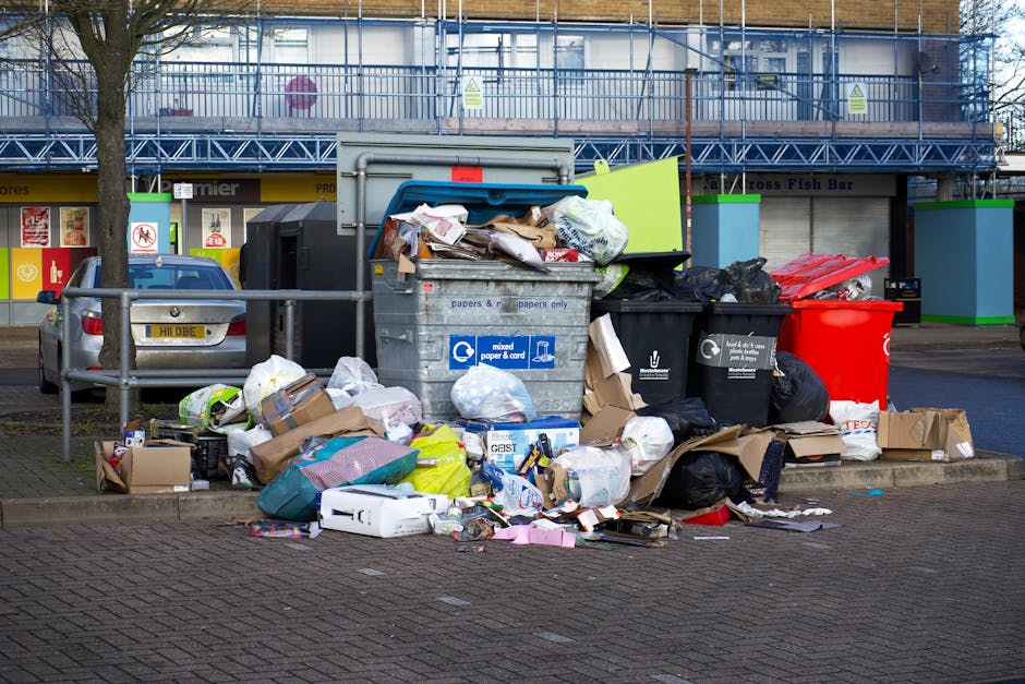 A collection of discarded household and outdoor items piled on a paved surface against a brick wall, including wooden garden furniture such as a slatted chair and a small table, both showing signs of