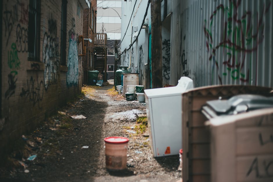 A collection of overflowing rubbish and waste bags piled around and inside a large grey mixed paper and cardboard recycling bin on a paved urban street. The waste includes cardboard boxes, plastic bag